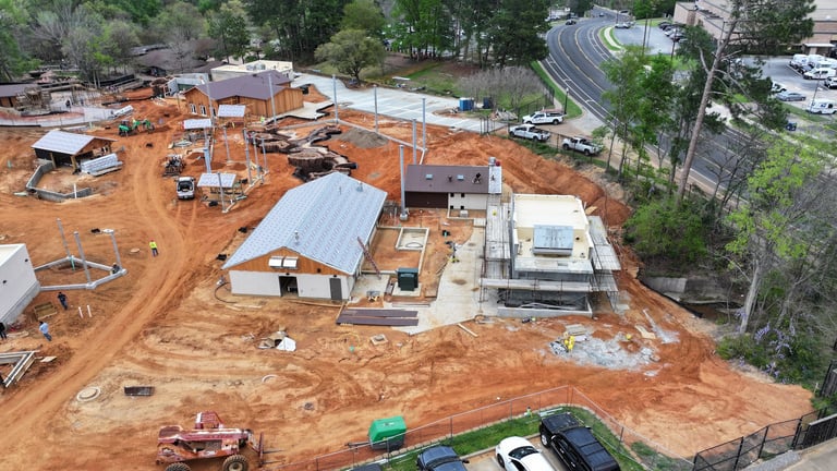 Aerial view of an active construction site with red clay soil, metal-roofed buildings under construction, and construction equipment scattered across the property