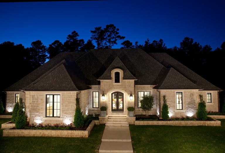 Modern brick home illuminated at night with stone exterior, arched entryway, manicured lawn, and tree-lined backdrop