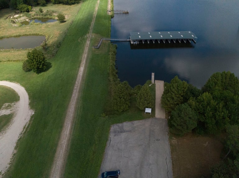 Aerial view of a boat dock and parking area next to a calm blue water body, with grassy areas and trees surrounding the waterfront facility