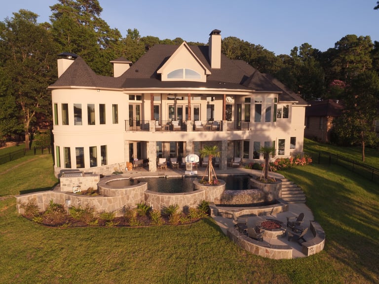 Large beige two-story luxury home with columned porch, stone patio, and manicured lawn surrounded by trees