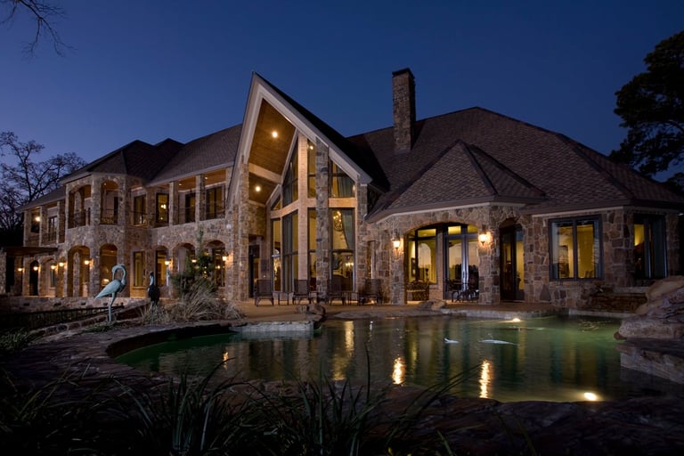 Luxury stone mansion illuminated at dusk with pool and landscape lighting reflecting on water