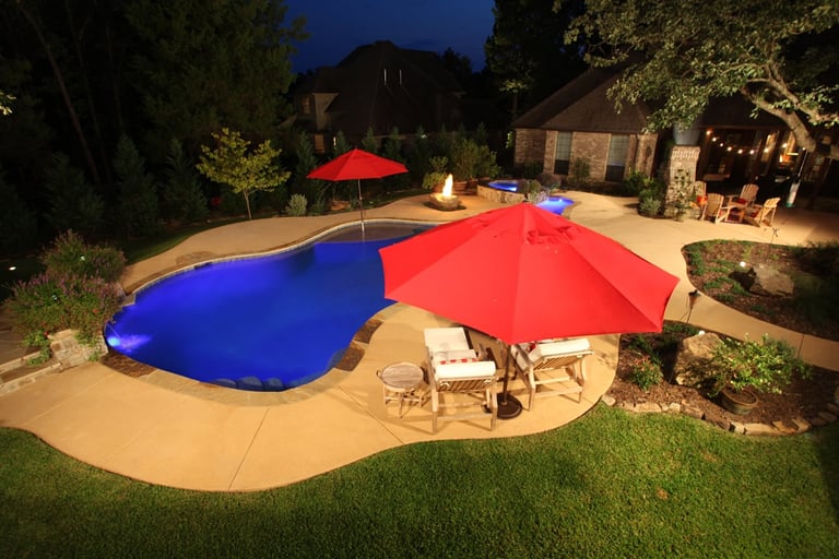 Nighttime backyard with an illuminated pool covered by blue and red pool covers, surrounded by concrete patio, chairs, umbrellas, and landscaping