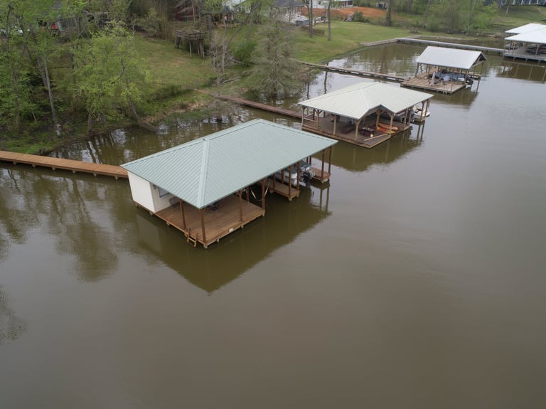 Aerial view of waterfront boat houses with metal roofs situated on a calm river surrounded by green trees