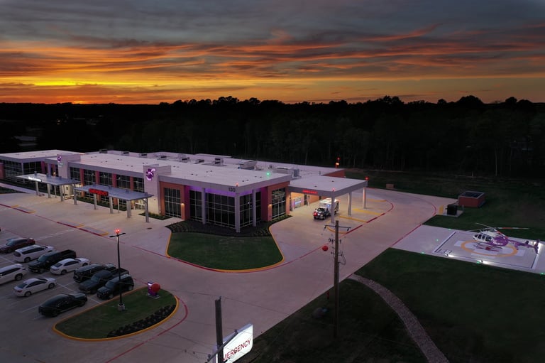 Modern commercial building with purple lighting at dusk, surrounded by parking lot and trees, dramatic orange sunset sky above