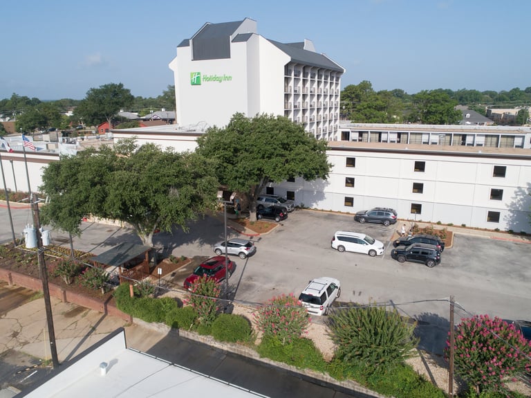 Holiday Inn hotel building with parking lot, trees, and landscaping viewed from above on a clear day