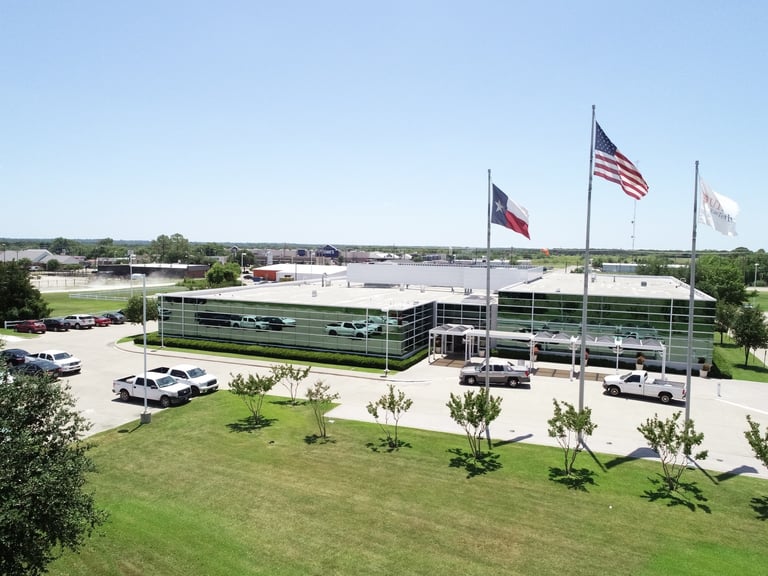 Modern commercial building with green glass facade, three flags, manicured grounds, parked vehicles, and landscaping