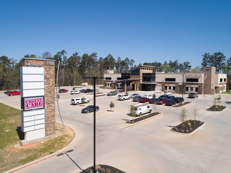 Aerial view of a commercial shopping center with brick buildings, parking lot with vehicles, and signage near a tree-lined perimeter