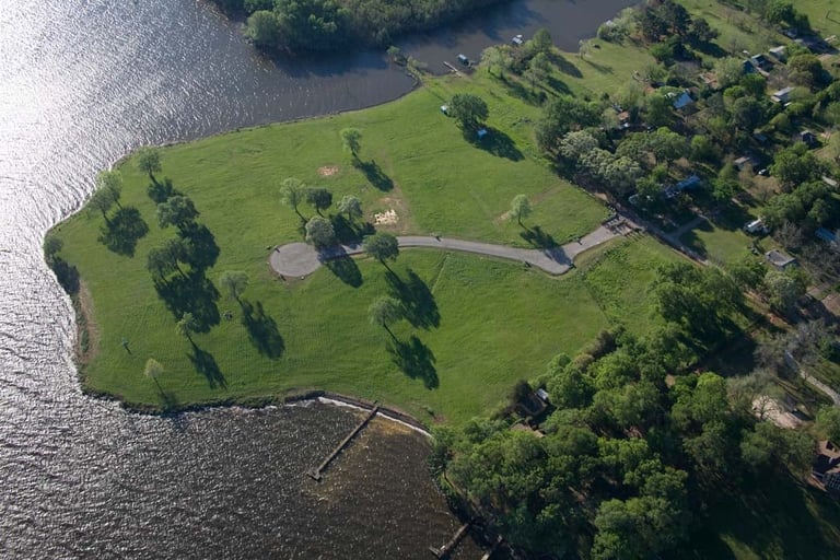 Aerial view of a green peninsula with a winding path, scattered trees, and a dock, surrounded by water