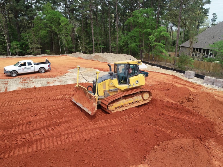 Yellow bulldozer grading red clay soil at construction site with white truck and tree line in background