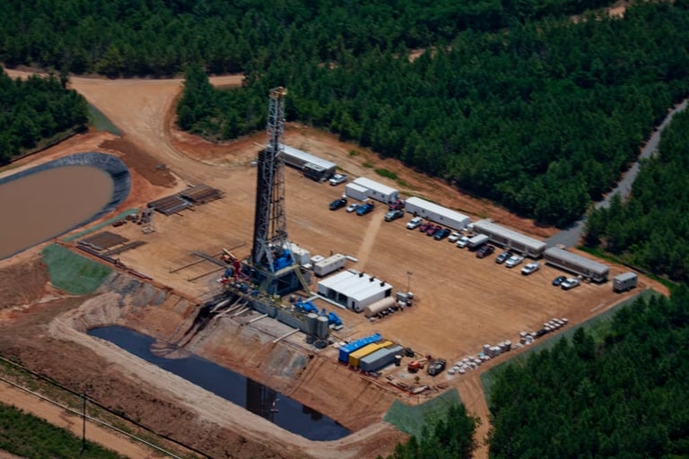 Aerial view of an oil drilling rig on cleared land surrounded by forest, with equipment and vehicles parked nearby