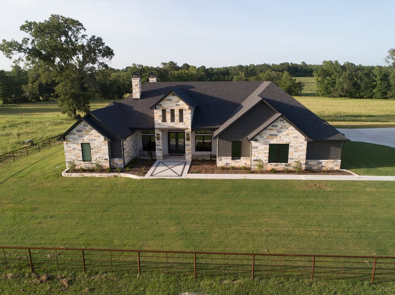 Modern farmhouse with dark roof and white stone facade on expansive green lawn bordered by metal fence, with trees and pond in background