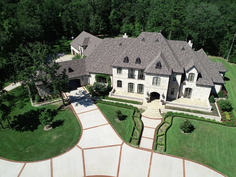 Aerial view of a large two-story stone mansion with slate roof, manicured grounds, circular driveway, and surrounding trees.