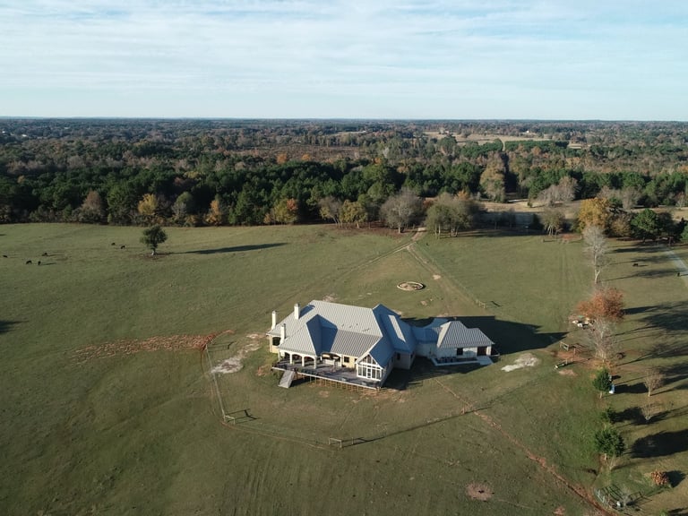 Aerial view of a large white farmhouse with a dark roof surrounded by green fields and trees in a rural landscape
