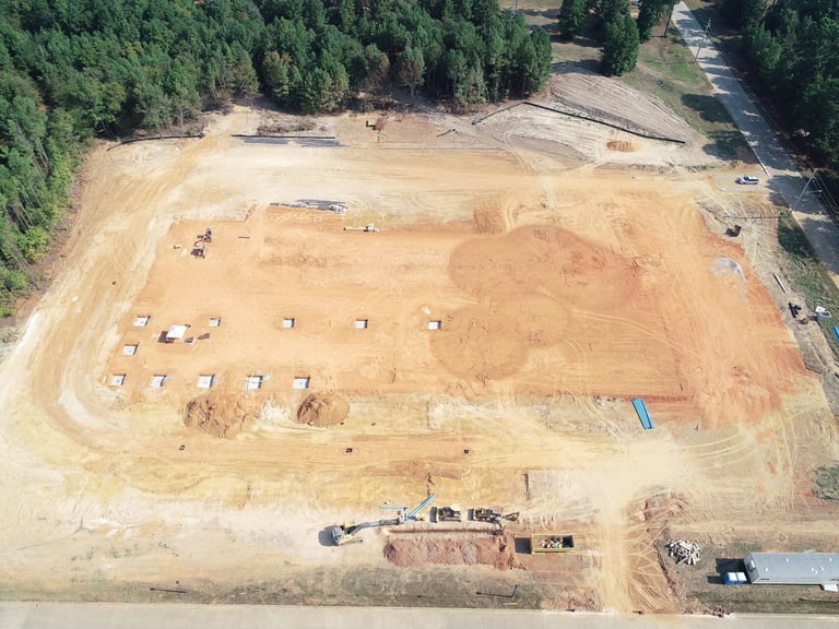 Aerial view of an active construction site with cleared land, equipment, and foundation work in progress surrounded by forest.