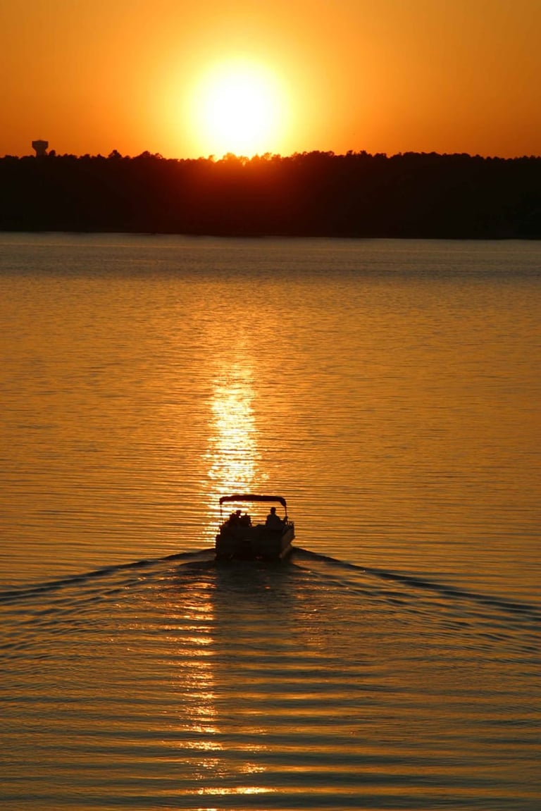 Small boat with passengers silhouetted on calm water during golden sunset with trees in background